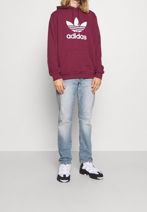 Vinröd luvtröja med vit Adidas-logga, kombinerad med ljusblå jeans och svartvita sneakers med mönstrade detaljer.