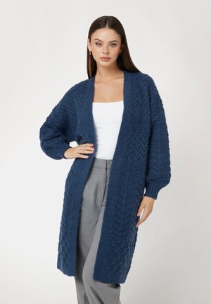 Dunkelblauer Strickcardigan mit strukturiertem Muster, offenem Vorderteil und langen Ärmeln. Über einem weißen Oberteil mit locker sitzenden grauen Hosen getragen.