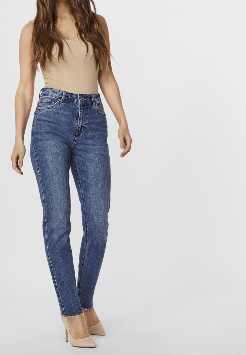 Femme portant un jean skinny bleu taille haute avec des ourlets bruts, un haut sans manches beige et des chaussures à talons beiges, se tenant devant un fond uni.