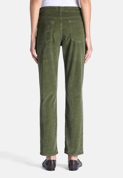 Personne portant un pantalon droit vert olive et des chaussures marron foncé, debout devant un fond clair uni, vue de dos.