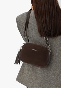 Borsa a tracolla in suede marrone con accenti in catena argentata, dotata di chiusura con zip e dettaglio con frangia. Logo impresso sul davanti.