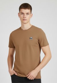 Camiseta de manga corta marrón, hecha de una tela suave, que presenta un pequeño gráfico de una cara con gafas de sol en el área izquierda del pecho.