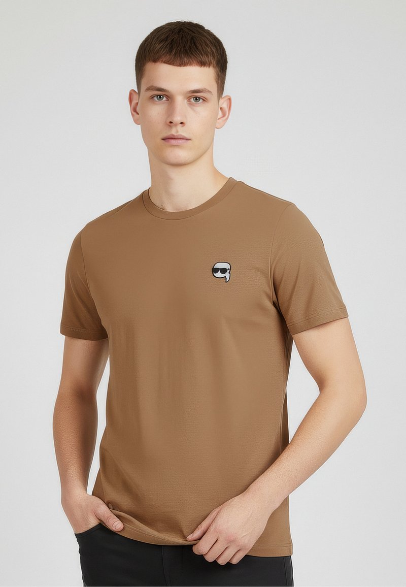 Braunes Kurzarm-T-Shirt aus glattem Stoff, mit einem kleinen Grafikmotiv eines Gesichts mit Sonnenbrille auf der linken Brustseite.