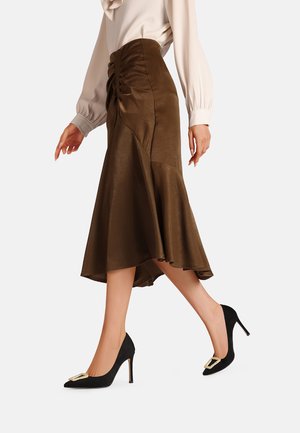 Gonna marrone al ginocchio con orlo svasato e arricciature laterali, abbinata a scarpe nere con tacco alto dotate di una fibbia decorativa.