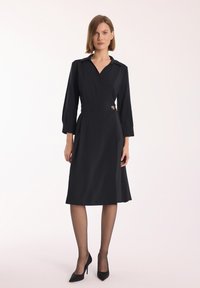 Czarna sukienka o kroju kopertowym z kołnierzykiem, rękawami o długości trzech ćwiartek i bocznym guzikiem. Gładka faktura, do kolan, z dopasowanym fasonem.