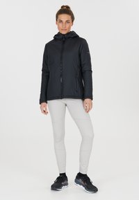 Veste isolante noire avec capuche, fermeture éclair à l'avant et poches latérales. Associée à des leggings gris clair et des chaussures de sport noires.