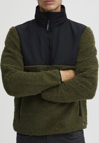 Fleece pullover with green textured fabric and a black smooth yoke, featuring a high collar, half-zip front, and side pockets.