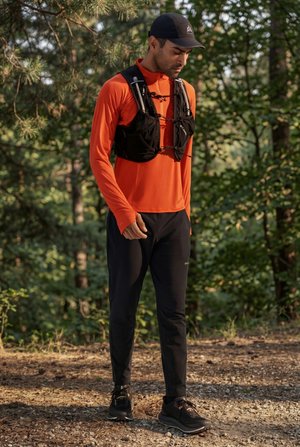 Mann i oransje langermet topp, svart løpevest, svarte bukser og caps, stående på en skogsti og ser ned.