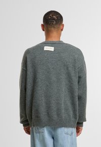 Siv volna pulover z ohlapnim krojem, rebrastim robom in pravokotno tkano oznako na hrbtni strani vratu, ki se ponaša z gladko teksturo.