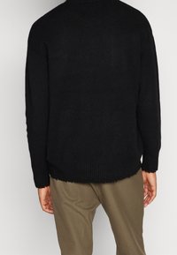 Svart stickad tröja med lös passform, med ribbat nederkant och ärmar. Kanterna är råa, vilket ger en texturerad detalj. Bärs över bruna byxor.