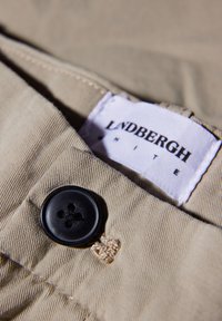 Tissu en coton beige avec un bouton noir à quatre trous et des coutures visibles ; une étiquette blanche indique "Lindbergh" en texte noir.