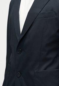 Σκούρο μπλε σακάκι με γιακά notch, δύο μαύρα κουμπιά και θήκες που φοριέται πάνω από λευκό πουκάμισο.
