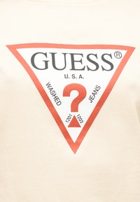 Μπεζ κοντομάνικο μπλουζάκι με μεγάλο κόκκινο τρίγωνο και το κείμενο «GUESS U.S.A.» καθώς και ένα ερωτηματικό. Στο κείμενο αναγράφονται οι λέξεις «WASHED» και «JEANS», με αριθμούς από κάτω.