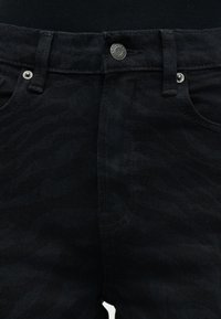 Κοντινό πλάνο μέσης μαύρου τζιν με θηλιές για ζώνη, εμπρόσθιες τσέπες και ασημέν ιο κουμπί κλεισίματος με ετικέτα επωνυμίας.