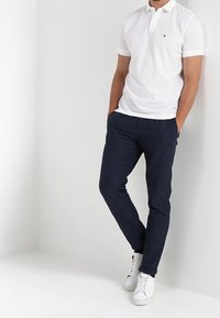 T-shirt polo blanc à manches courtes, associé à un pantalon de sport bleu marine extensible et des baskets blanches. Logo notable sur la poitrine du t-shirt.