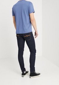 Blå t-shirt med kort ärm, mörk denimjeans med orange sömnadsdetaljer, slim fit, kombinerad med svarta högskaftade sneakers.