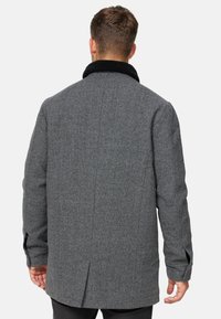 Abrigo de lana gris con cuello de piel sintética negra, con corte recto, costuras laterales y mangas largas, con detalle de abertura en la parte posterior.