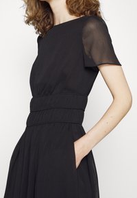 Černé šaty s přiléhavým tělem a skládací sukní, mají krátké průhledné rukávy a nařasený pas. Součástí jsou boční kapsy.