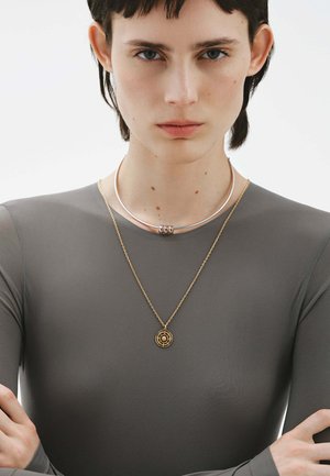 Collares de plata y oro superpuestos sobre una camiseta de manga larga gris. El chokers de plata presenta tres anillos metálicos; el colgante de oro tiene un diseño circular.