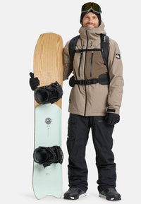 Snowboard features a wooden top with a mint green base, paired with black bindings. The rider wears a light brown jacket and black pants.