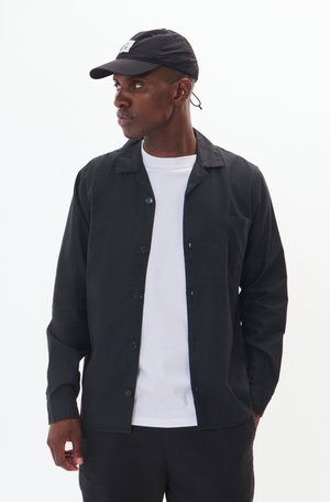 Black button-up shirt with a pocket, worn over a white t-shirt. The model wears a black cap with a visible logo. Plain background.