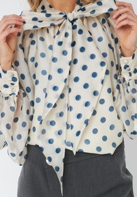 Camicia bianca con pois blu, dotata di un grande fiocco al collo e maniche a palloncino con polsini abbottonati. Tessuto morbido e leggero.