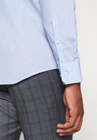 Μανσέτα γαλάζιου πουκαμίσου με μακριά μανίκια και κουμπί στερεωμένο πάνω σε γκρι καρό παντελόνι, με ένα χαλαρό χέρι στο πλάι.