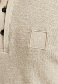 Beige strukturierte Hemd mit Kragen, drei schwarzen Knöpfen und einem quadratischen Logo-Patch. Der Stoff wirkt weich und leicht gewebt.