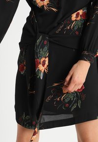 Vestido negro con estampado floral y mangas largas, que presenta vibrantes patrones de flores naranjas y rojas, un detalle de cintura atada y una abertura lateral.