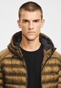 Brązowa puchowa kurtka z kapturem, charakteryzująca się pikowaną teksturą i czarną podszewką. Nosi ją model na czarnej koszuli, zbliżenie na twarz modela.