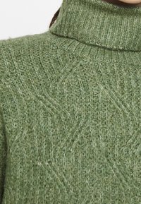 Un pull col roulé en tricot vert avec un motif en vagues texturé. Tissu doux et côtelé avec un col haut et des détails de couture visibles.