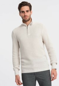Pull en tricot beige clair avec un col zippé quart, motif texturé, poignets et ourlet côtelés. Porté sur un pantalon gris.