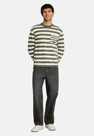 Man glimlacht, draagt een groen-wit gestreept shirt met lange mouwen en zakje, donkere spijkerbroek en witte sneakers, staand tegen een neutrale achtergrond.