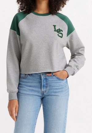 Kobieta ubrana w krótki szary sweter z zielonymi ramionami i haftowanym logo LS, w połączeniu z jasnoniebieskimi jeansami z wysokim stanem.