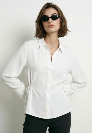 Femme aux cheveux courts et bruns portant des lunettes de soleil ovales noires, une chemise blanche boutonnée avec des liens sur les côtés, et un pantalon noir, debout sur un fond blanc.