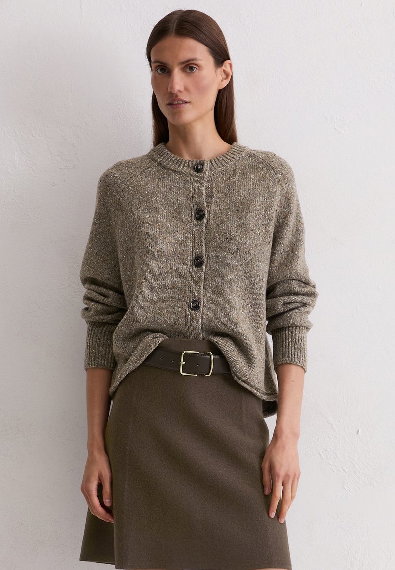 Frau in einem braunen, knöpfbaren Strickpullover, der in einen passenden braunen Rock mit einem schwarzen Gürtel gesteckt ist, steht vor einer schlichten weißen Wand.