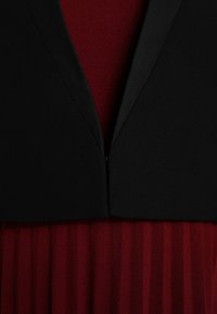 Top negro con cuello tipo polo y escote en forma de V sobre un vestido rojo texturizado, con pliegues verticales que crean una apariencia de capas.