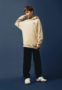 Sudadera con capucha beige con gráfico "NYC Brooklyn", combinada con pantalones negros y zapatillas blancas. Tejido suave y corte relajado.