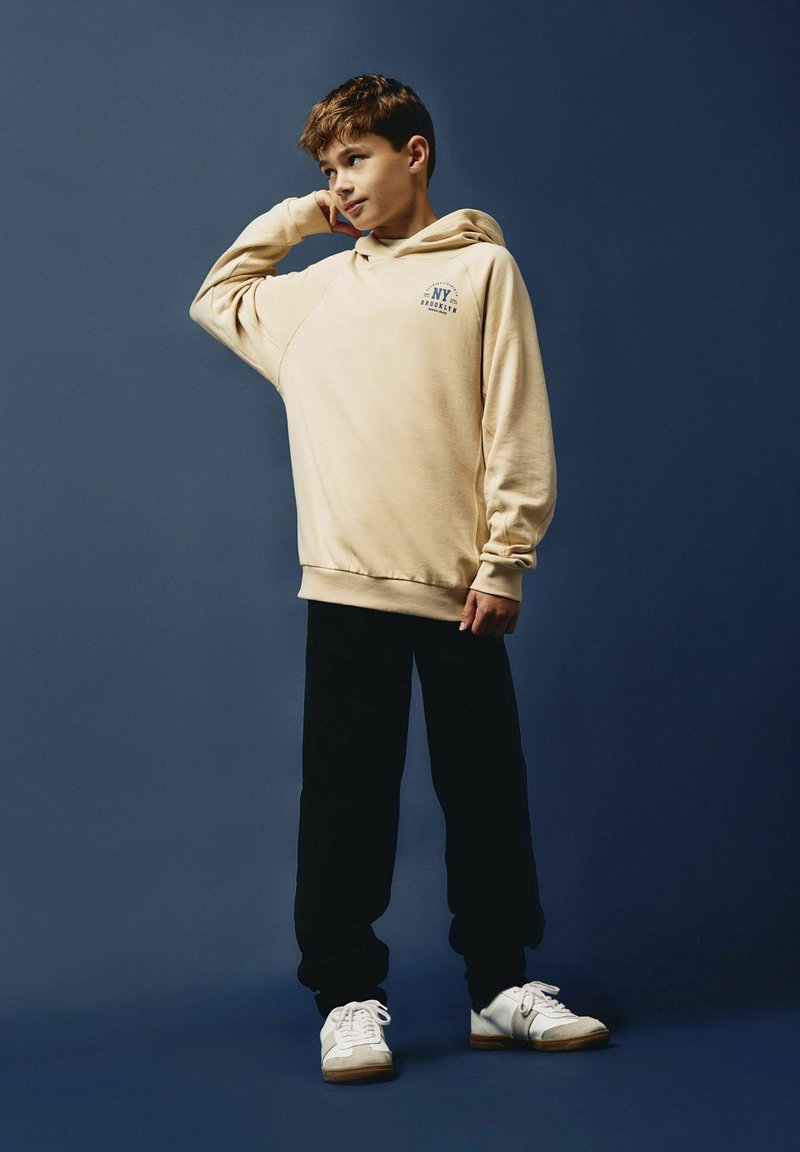 Sudadera con capucha beige con gráfico "NYC Brooklyn", combinada con pantalones negros y zapatillas blancas. Tejido suave y corte relajado.