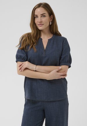 Femme aux longs cheveux châtain clair, portant un haut à manches courtes texturé bleu marine et un pantalon assorti, debout les bras croisés devant un fond blanc.