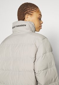 Světlá šedá péřová bunda s vysokým límcem a černým logem "HELLO HANSEN". Obsahuje prošívaný design a mírný lesk.