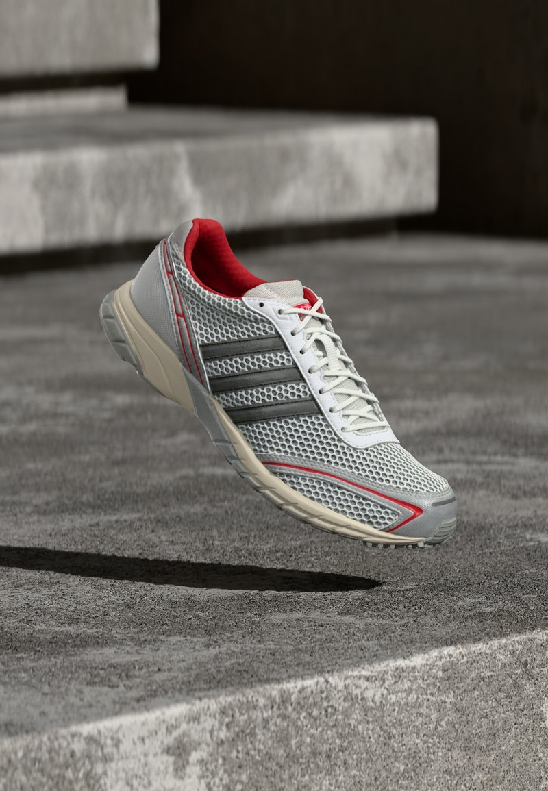 Chaussure de sport grise avec une tige en mesh, une doublure intérieure rouge, des lacets blancs et trois bandes noires. Semelle gris clair avec des accents rouges.