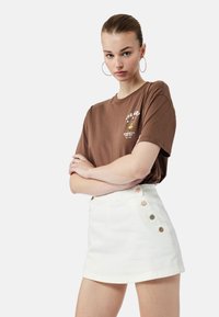 Camiseta de algodón marrón con estampado gráfico, combinada con una minifalda de mezclilla blanca con detalles de botones laterales y una cintura ajustada.