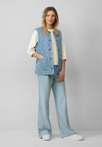 Gilet matelassé bleu clair avec un devant à boutons, associé à une chemise blanche à rayures et un pantalon flared en denim clair. Il comporte des poches latérales.