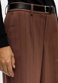Pantalons marron à rayures fines en tissu doux, dotés d'une ceinture en cuir noir et d'une coupe ajustée. Poches avant visibles et coutures soignées.