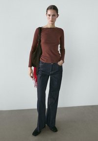 Kvinde står op ad grå væg iført brun langærmet top, mørke bukser med vide ben, sorte sko og bærer en sort taske med et rødt tørklæde.