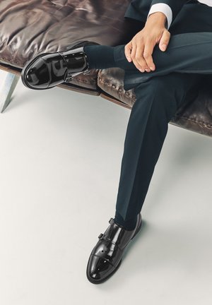 Hombre con traje oscuro sentado con las piernas cruzadas en un banco de cuero marrón, llevando zapatos de vestir negros brillantes y calcetines azul marino.