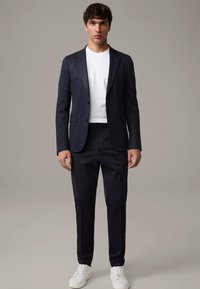 Granatowa marynarka garniturowa z dopasowanymi spodniami i biała koszulka, połączone z białymi trampkami. Krój na miarę, teksturowana tkanina, czyste linie.
