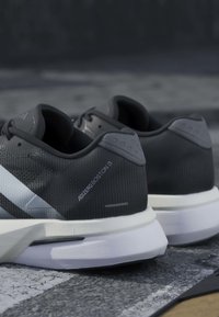 Paire de chaussures de course Adidas Adizero Boston 13 en noir et blanc posée sur une surface extérieure texturée.