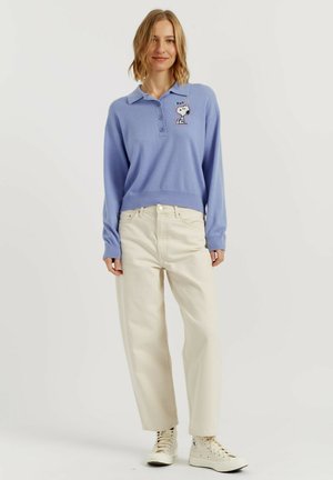 Femme portant un pull polo à manches longues violet clair avec broderie Snoopy, pantalon large crème et baskets montantes blanches.